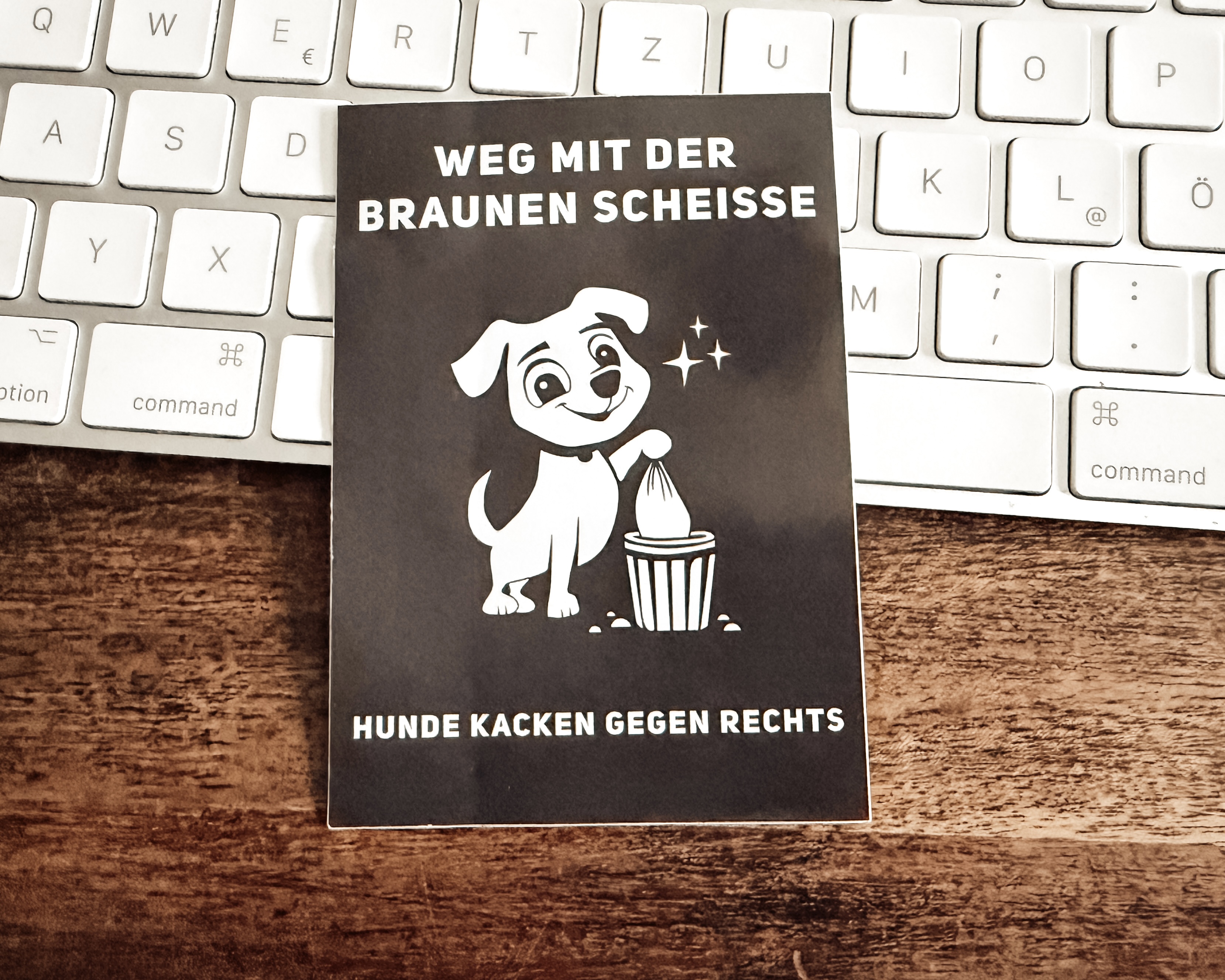 Ein Aufkleber mit der Aufschrift „Weg mit der braunen Scheiße“ und der Illustration eines Hundes, der einen Beutel in einen Mülleimer wirft, lehnt an einer Tastatur auf einem Schreibtisch 
