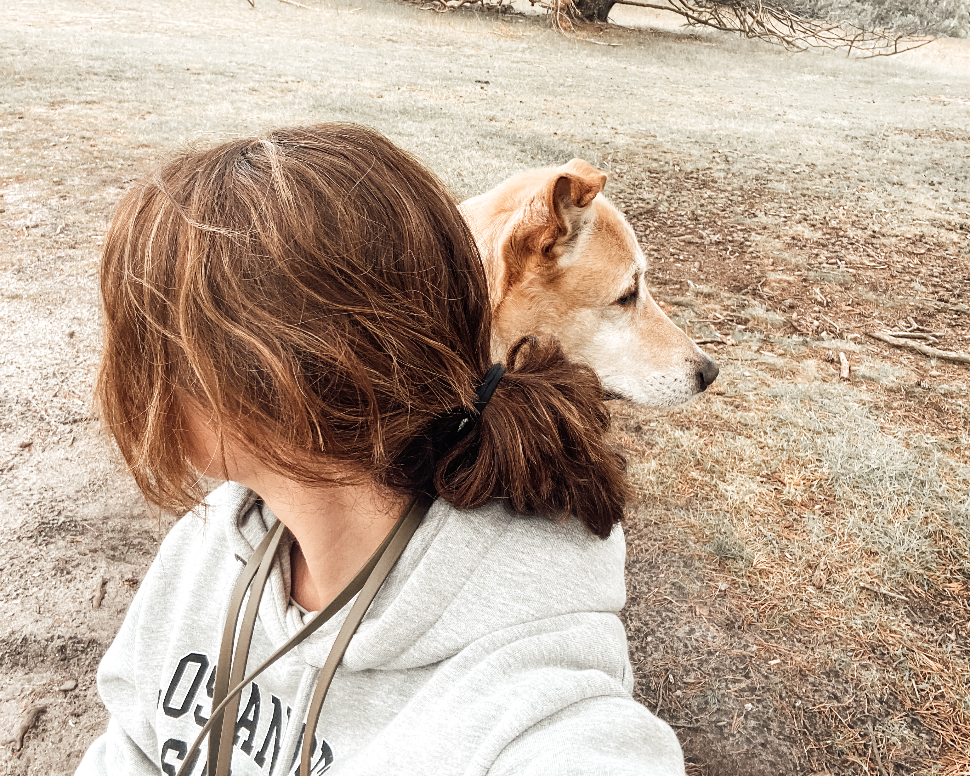 Eine Frau sitzt mit ihrem Hund in offener Landschaft. Beide schauen in unterschiedliche Richtungen.